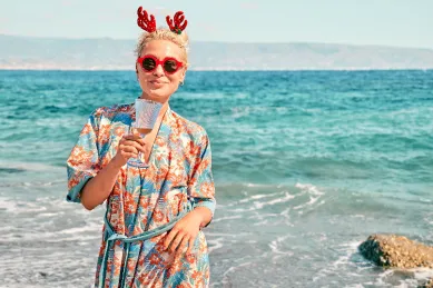 Junge Frau mit blonden Haaren, rotem Rentiergeweih-Haarreif und Sonnenbrille stößt mit einem Sektglas an einem sonnigen Strand an, im Hintergrund das türkisblaue Meer. Symbolbild für die Auszeit die Ihnen die AIDA Winterpause ermöglicht.