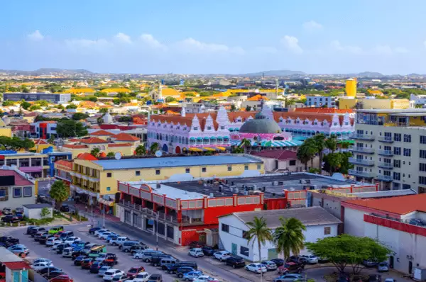 Schon vom Kreuzfahrtschiff aus können Sie einen Blick auf die Shoppingmöglichkeiten in Oranjestad werfen. Blick von einem Kreuzfahrtschiff aus auf einen Parkplatz, Einkaufszentrum und Geschäfte in Oranjestad, Aruba.