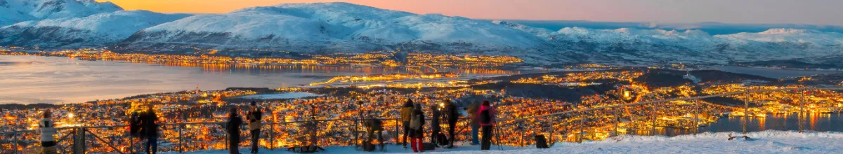 Weitwinkel-Aufnahme vom Aussichtspunkt über das nächtlich beleuchtete Tromsø in Norwegen mit Touristen an der Brüstung während einer Nordland-Kreuzfahrt mit der MSC Preziosa.