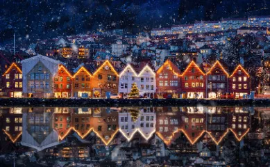 CATCH OF THE WEEK: Nordeuropa mit der MSC Schiffvielfalt Eine nächtliche Winteraufnahme der historischen Bryggen-Häuser in Bergen, Norwegen, bei leichtem Schneefall. Die farbenfrohen Giebelhäuser sind festlich mit goldenen Lichterketten an den Dachkanten beleuchtet und spiegeln sich auf dem Wasser.