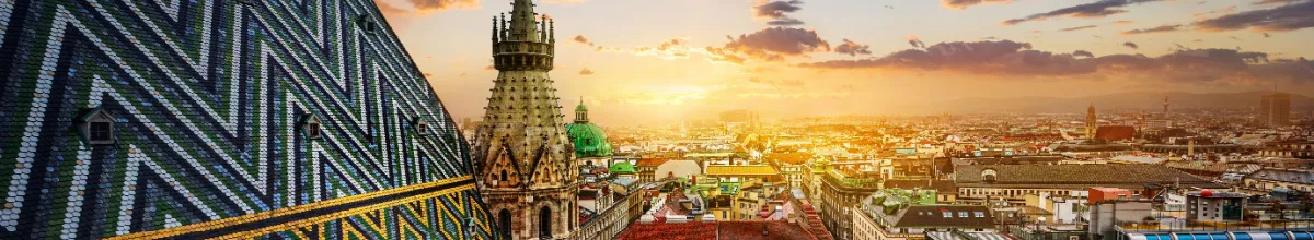 Das charakteristisch bunt gemusterte Ziegeldach und die steinernen Türme des Stephansdoms über den Dächern von Wien im warmen Licht des Sonnenuntergangs.