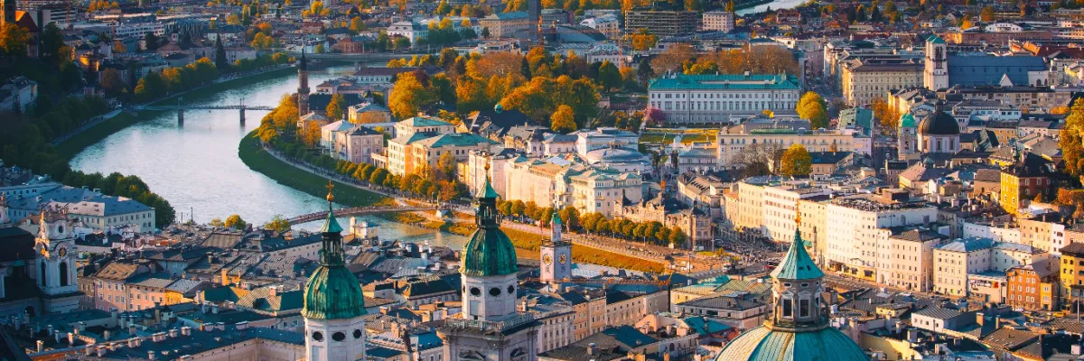 Blick von oben auf die barocke Altstadt von Salzburg mit den grünen Kuppeln des Doms und der geschwungenen Salzach im warmen Abendlicht.