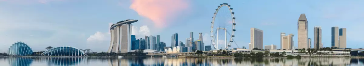 Skyline von Singapur an der Marina Bay mit dem geschwungenen Gewächshaus Flower Dome, dem Hotel Marina Bay Sands und dem Riesenrad Singapore Flyer. Die Gebäude spiegeln sich im Licht der Dämmerung in den blauen Fluten des Südchinesischen Meers.