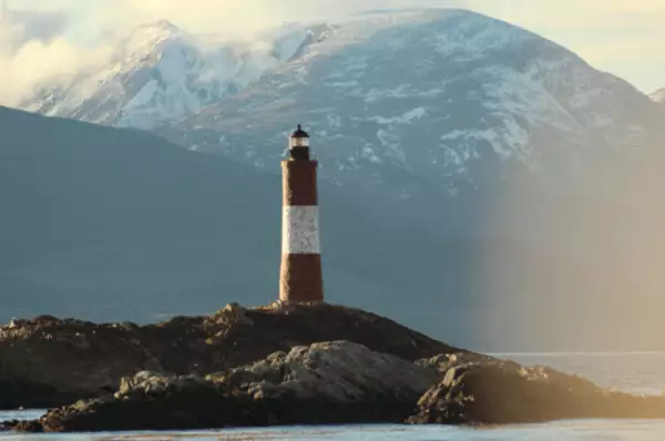 Der rot-weiße Leuchtturm steht auf einer kleinen Felsinsel im Beagle-Kanal vor einem Berg mit schneebedeckten Hängen.