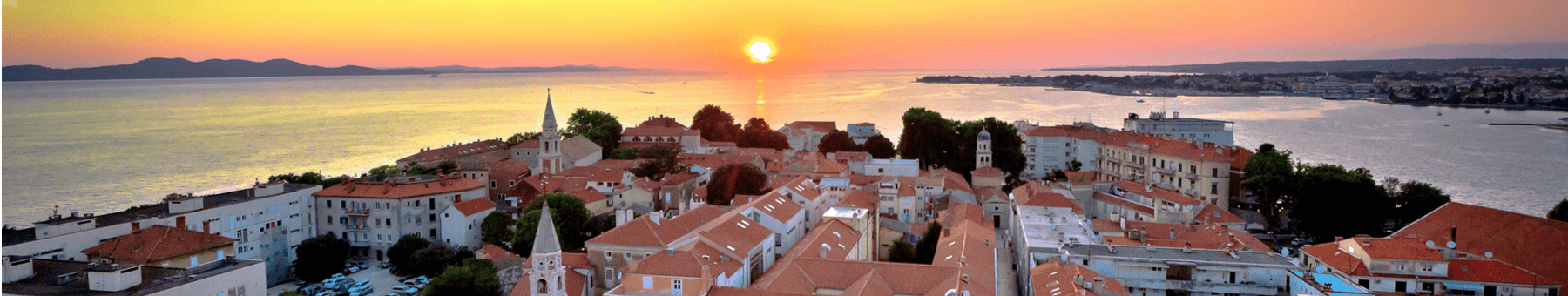 Im Hintergrund geht die Sonne unter. Demnach schimmert der Himmel rötlich. Davor ist das Meer welches hinter der Stadt hervorragt. Die Stradt erstrahlt im Sonnenlicht.