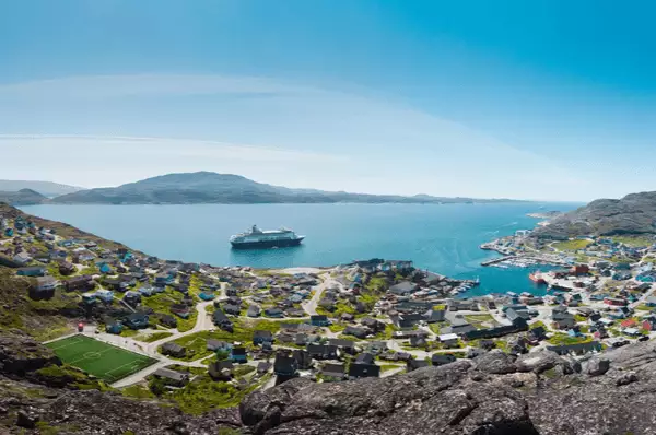 Eine Panoramaaufnahme der grönländischen Küste, Qaqortoq, mit einem Kreuzfahrtschiff auf dem Meer mit blauem Himmel und kleinen bunten Holzhäusern