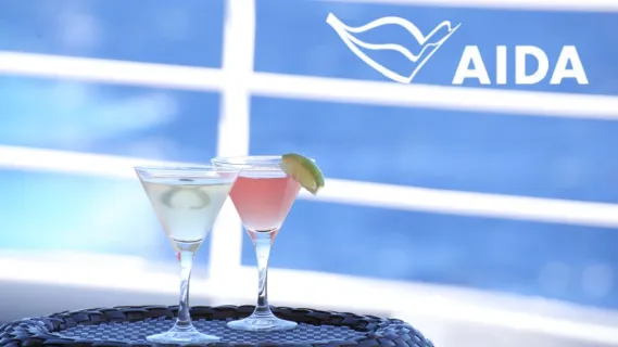 Mit AIDA All Inclusive zu den schönsten Zielen der Welt 2 Cocktailgläser stehen auf einem dunklen runden Tablett vor der weißen Reling eines Kreuzfahrtschiffes. In der rechten oberen Ecke ist das AIDA Logo in weiß zu sehen.
