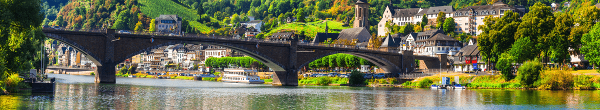 Eine mittelalterliche Brücke, die sich über die Mosel spannt, mit der mittelalterlichen Stadt Cochem dahinter. Im Hintergrund sind üppige Wälder.