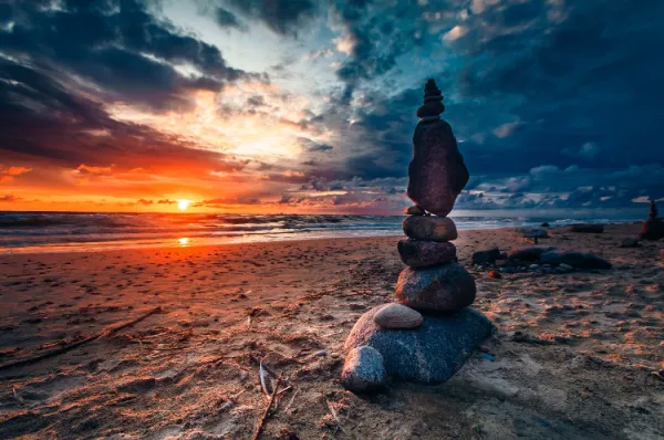 Ein Turm aus gestapelten Steinen an einem Sandstrand, im Hintergrund ein dramatischer Sonnenuntergang über dem Meer mit farbenfrohen Wolken.