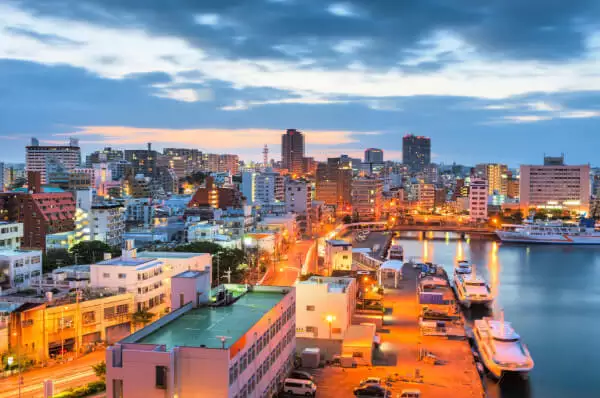 Naha Hafen und Innenstadt bei Morgendämmerung mit Häuserskyline, erleuchteten Lichtern und ankernden Booten, Okinawa, Japan