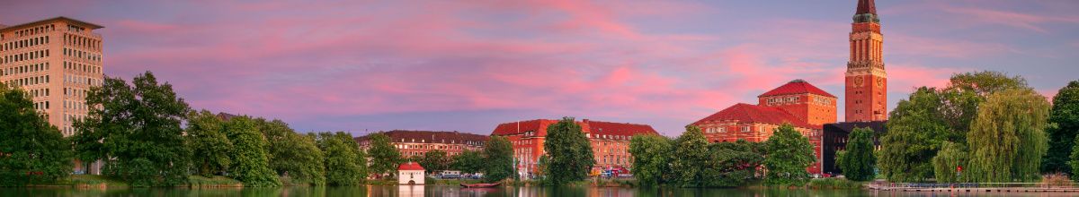 Panorama des Kleinen Kiels mit dem markanten Rathausturm und dem Opernhaus Kiel vor einem farbenprächtigen, rosa leuchtenden Abendhimmel.