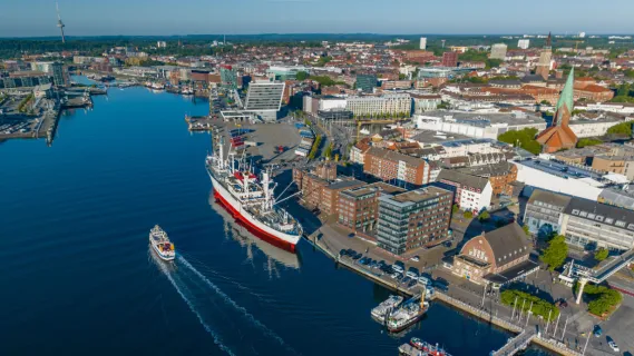 Luftaufnahme der Kieler Förde und der Hörn mit einer fahrenden Fördefähre, einem historischen Schiff am Kai und der Kieler Innenstadt im strahlenden Sonnenlicht.
