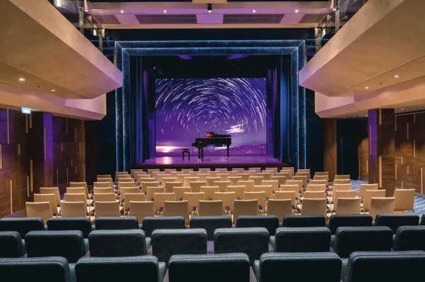 Eine Innenaufnahme des Theatersaals an Bord des Kreuzfahrtschiffs <i>Mein Schiff 7</i>. Der Saal zeigt Reihen von blauen und beigen Sitzen, die auf eine Bühne ausgerichtet sind. Auf der Bühne steht ein schwarzer Flügel vor einer großen digitalen Leinwand.