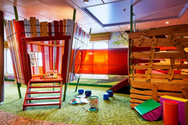 Eine Innenaufnahme eines farbenfrohen Kinderspielbereichs an Bord der Mein Schiff 7. Der Raum ist mit einer hölzernen Kletterstruktur ausgestattet, die über eine kleine rote Treppe und eine große rote Röhrenrutsche verfügt.