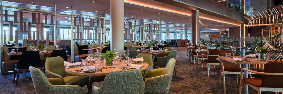 Eine Panoramaaufnahme eines der modernen Restaurants auf dem Kreuzfahrtschiff <i>Mein Schiff 7</i>. Der stilvoll eingerichtete Speisesaal verfügt über runde Holztische, die mit Geschirr und Gläsern eingedeckt sind, umgeben von bequemen grünen Stühlen.