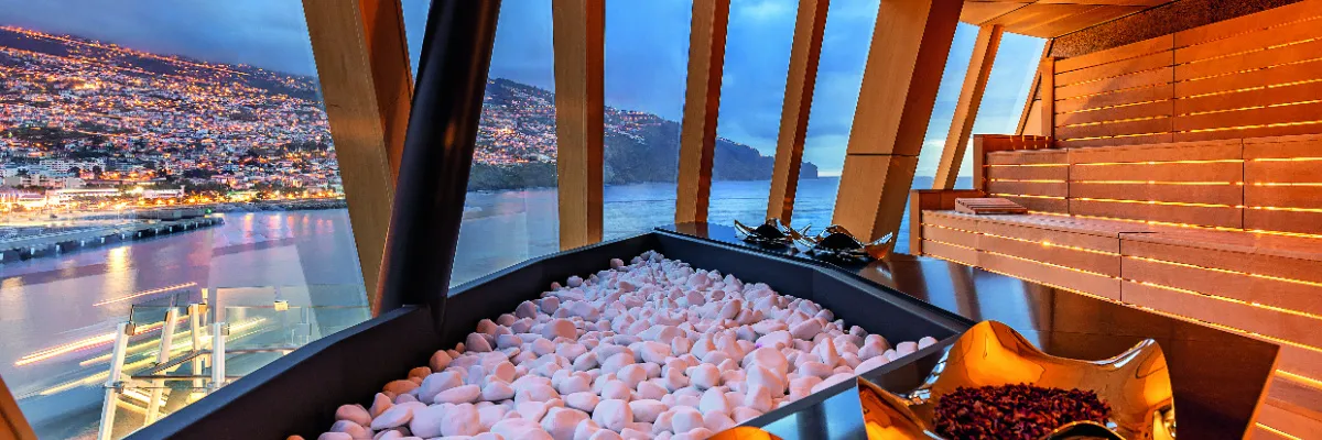 Eine Panoramaaufnahme aus dem Spa-Bereich der <i>Mein Schiff 7</i>, die eine holzverkleidete Sauna mit großen, schrägen Fenstern zeigt. Durch die Fenster blickt man auf eine erleuchtete Küstenstadt und einen Hafen bei Dämmerung.