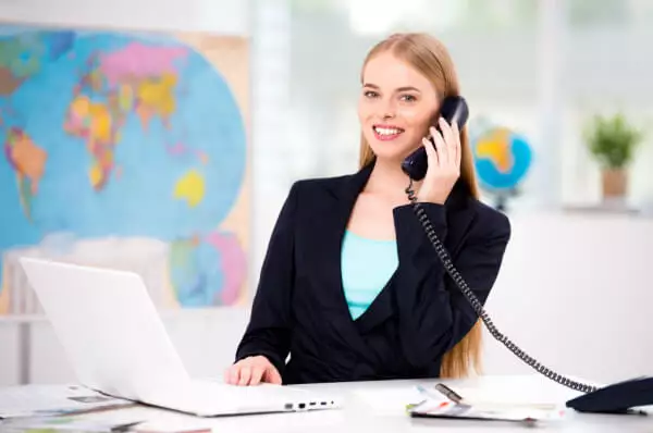 Eine junge Frau sitzt telefonierend mit einem Laptop vor einer Weltkarte.