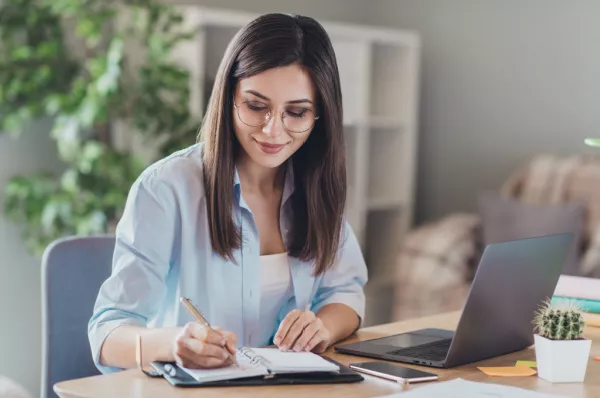 Eine junge Frau sitzt am Schreibtisch und schreibt etwas in ihr Notizbuch. Daneben sind ein rosa Smartphone und ein aufgeklapptes Notebook zu sehen.