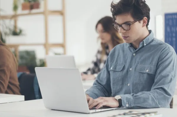 Junger Mann an einem weißen Laptop, im Hintergrund zwei junge Frauen, ebenfalls an Laptops.