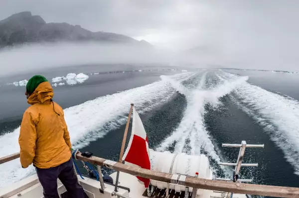 Ausblick beim Einlaufen auf den Hafen Eine Person mit gelber Jacke und grüner Mütze steht hinten auf dem Schiff und blickt in Richtung Hafen von Nuuk. Hinter dem Boot im Wasser sieht man die Wellen, die der Motor schlägt.