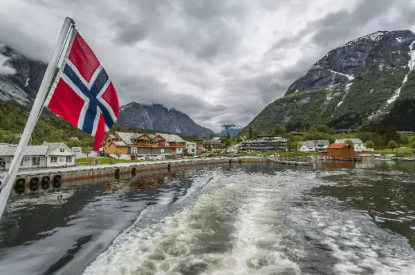Wenn man hinten auf einem Boot steht, sieht man dieses Bild. Die Spuren die das Boot im Wasser hinterlässt, die norwegische Flagge welche hinten auf dem Boot hängt, sowie die Gebirgskette und das Dorf, von welchem Sie sich entfernt.