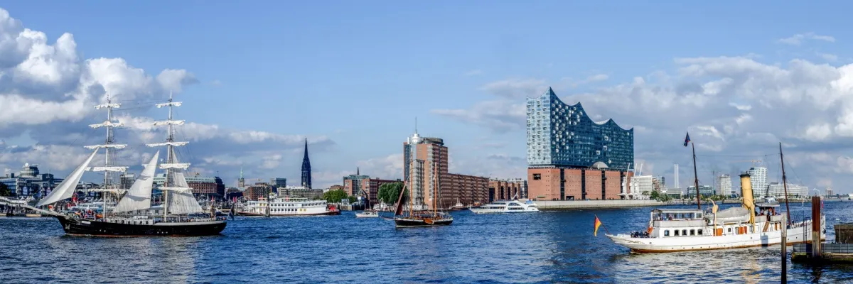 Panoramaansicht des Hamburger Hafens. Im Vordergrund zwei Segelboote und einige Ausflugsschiffe auf der Norderelbe, im Hintergrund die Elbphilharmonie und ein Kirchturm.