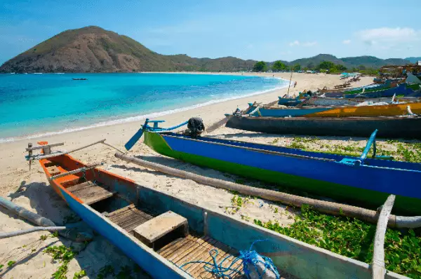 Boote am Strand Auf weißem Sandstrand liegen kleinere Boote im Vordergrund des Bildes. Dahinter ist das türkisblaue Wasser zu sehen, über dem sich im Hintergrund ein Vulkan erhebt..
