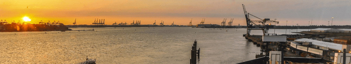 Rechts im Bild sind Teile des Hafens zu sehen. Links geht die Sonne unter. Mittig ein Fluss, welcher im Hafen mündet