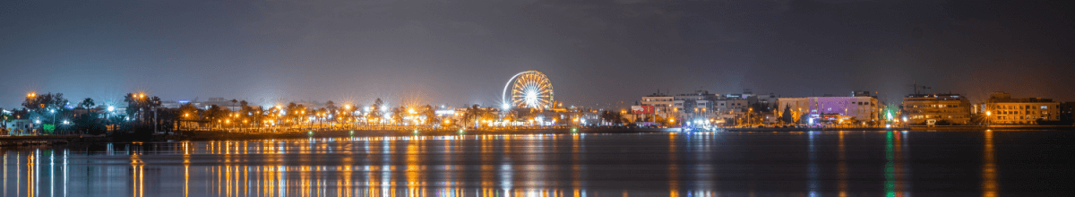 Das Panorama zeigt Tunis bei Nacht. Neben einem Riesenrad sind über das Gewässer eine Vielzahl an hellen, bunten Lichtern zu sehen.