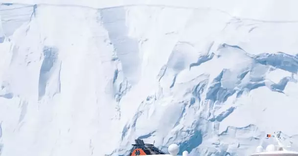 Am Ufer laufen Pinguine übers Eis. Übers Meer fährt die HANSEATIC spirit vor einem Eisberg.