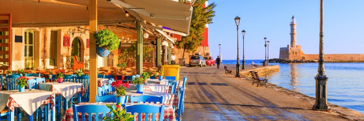 Malerische Szene eines Restaurants mit blau-weißen Tischen und Stühlen auf einer Terrasse direkt an einem Hafen im Mittelmeer. Im Hintergrund ist ein historischer Leuchtturm an der Hafeneinfahrt zu sehen.