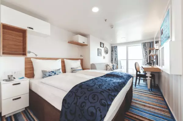 Ihr Wohlfühl-Refugium: Balkonkabine auf Mein Schiff 3 Blick in eine Balkonkabine auf Mein Schiff 3. Doppelbett mit blauer Tagesdecke, ein blauer Fußboden und ein weißer Nachttisch. Im Hintergrund ein Stuhl und ein Schreibtisch und die Glastür zum Balkon.