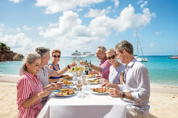 Glückliche Gäste beim Gourmet-Essen am Strand mit Blick auf ein Hapag-Lloyd Cruises Schiff, symbolisch für den Luxus im Platin-Tarif.