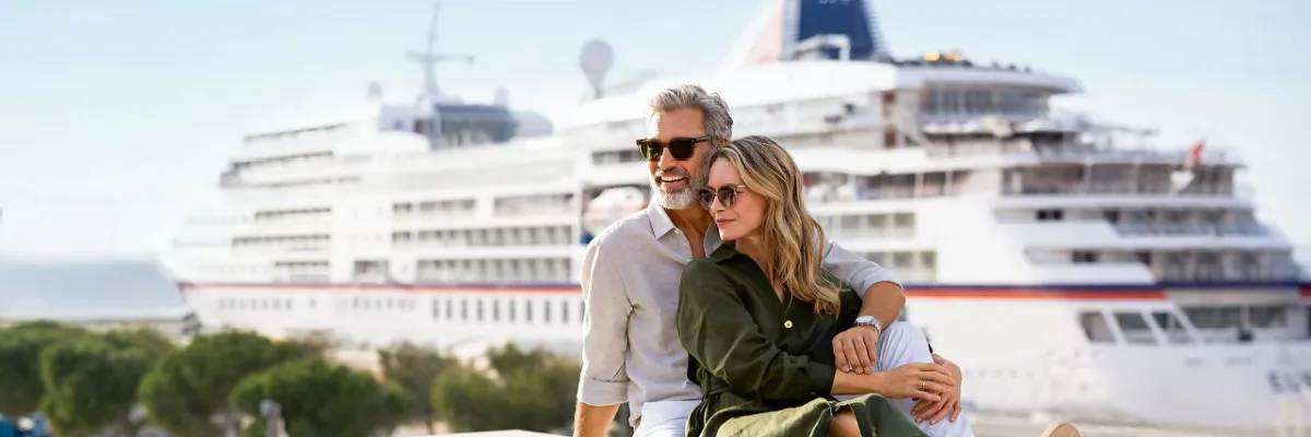 Ein glückliches Paar sitzt entspannt an der Hafenpromenade, im Hintergrund liegt ein weißes Hapag-Lloyd Kreuzfahrtschiff vor Anker – symbolisch für die Vorfreude auf die Reise.
