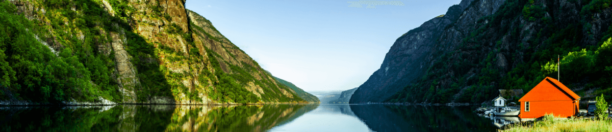 Ein kleines rotes Holzhaus steht an einem norwegischen Fjord mit hohen Felswänden.
