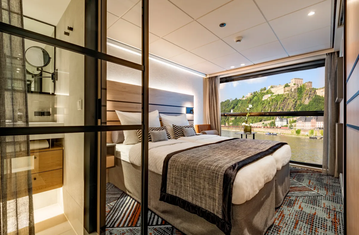 Elegante Außenkabine der nickoSPIRIT mit Doppelbett, stilvollem Bad und elektrisch absenkbarem Panoramafenster mit Blick auf das Rheintal