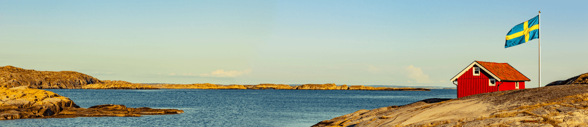 Rotes Holzhaus am Wasser an der schwedischen Küste. Daneben die Flagge Schwedens.