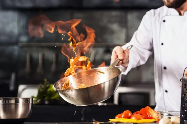 Ein Sternekoch in Schürze wendet Essen in einem Wok, der Flammen aufsteigen lässt. Live zum Miterleben während einer Eventkreuzfahrt mit nicko cruises.