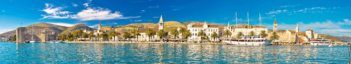 Panoramablick auf die Hafenstadt Trogir, auf die Uferpromenade mit Palmen und Schiffen davor.