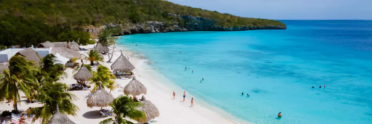 Sonnenschirme an einem weißen Sandstrand auf Curacao, rechts daneben das azurblaue Meer. Im Hintergrund eine dicht bewaldete Landzunge.