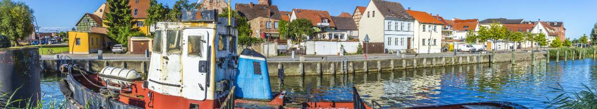 Die Uferpromenade in Wolgast, im Vordergrund liegt ein altes Schiff und im Hintergrund stehen Häuserreihen.