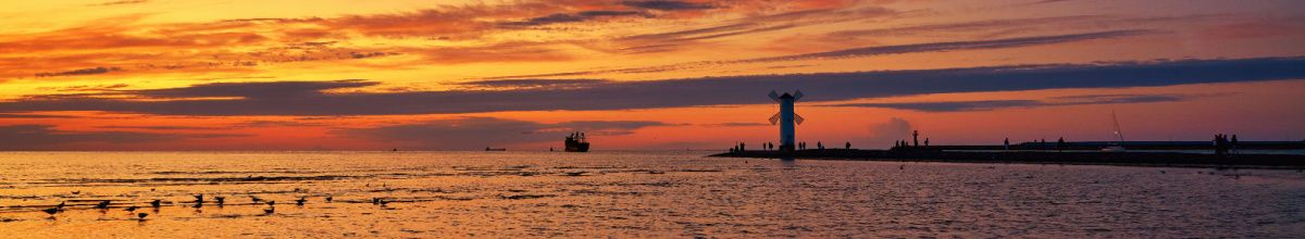 Die Küste von Swinemünde bei Sonnenuntergang mit Silhoutten eines Leuchtturmes und Schiffs in der Ferne während der Himmel vom Abendfeuer geküsst wird.