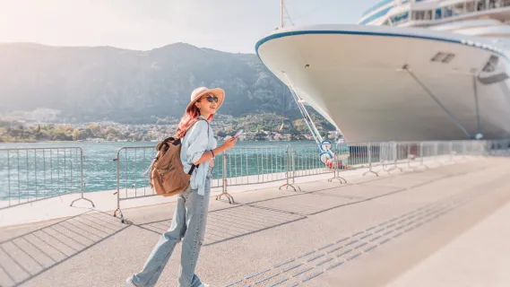 Zum ersten Mal auf einem Kreuzfahrtschiff: Tipps & Tricks Eine junge Frau mit Sonnenhut und Rucksack blickt am Hafen auf ein großes Kreuzfahrtschiff.